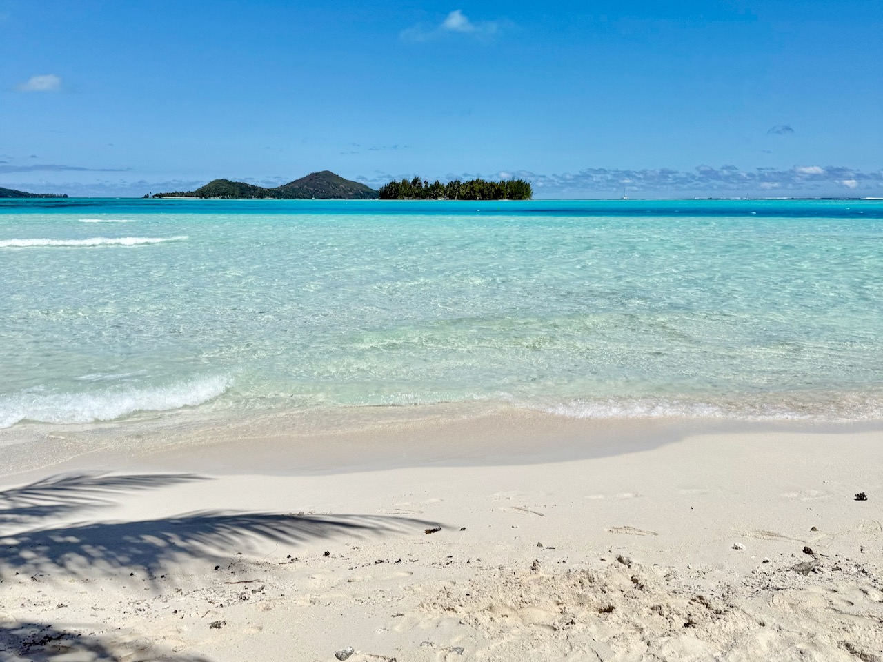 Quiet, empty beach inside the resort — palm shadow on white sand, distant motu across the lagoon